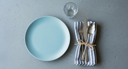A clean and fresh place setting on a textured gray background. The flat lay composition features a smooth, light blue plate and a bundle of rustic silver cutlery wrapped in a blue-and-white striped na