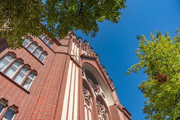 A red-brick church facade rises against a clear blue sky. Green trees frame the historic building...