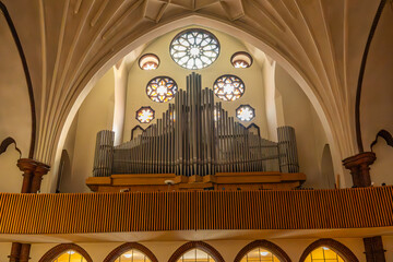 A large pipe organ beneath round stained-glass windows dominates the church balcony. Light shines...