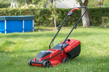 A red lawn mower stands on green grass in a backyard. In the background, a blue pool and hedge are...