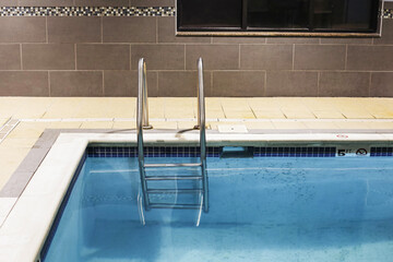 Indoor swimming pool with clear blue water and stainless steel ladder. Empty modern spa and fitness facility. Relaxation and leisure background. Clean tiled edge of poolside. Tranquil wellness concept