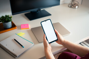A person is engaged in work from home, holding a smartphone with a blank screen in hands. The tidy...