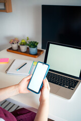 A person holds a smartphone while seated at a clean desk with a laptop, notebook, and plants....