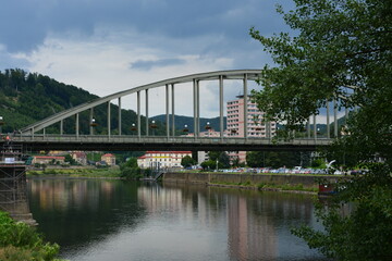 A large steel arch bridge spans a wide, calm river, connecting the two sides of a town. A dramatic,...