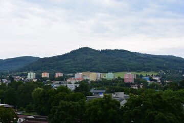 Colorful pastel apartment buildings stand in a valley at the base of a vast, densely forested mountain under an overcast sky