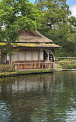Obraz premium Serene japanese watermill house by pond with lush greenery in peaceful rural setting concept of travel agencies, architecture firms, nature retreats in Japan
