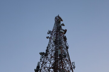 A towering metal telecommunications mast with antennas stands against a blue sky, connecting...