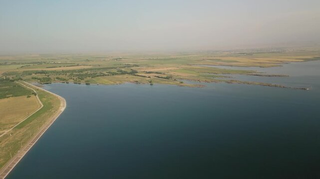 Aerial view of Djizak Reservoir dam and adjacent farmlands. Fields stretch toward the horizon. Water meets rural roads and agricultural land under bright, expansive skies in Uzbekistan