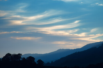 Obraz premium silvery clouds touching treetops of layered hills under evening light