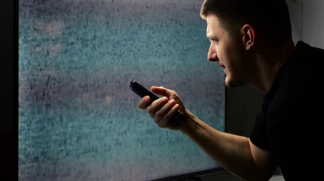 A young guy in front of a TV screen can't tune the channel and sees interference and white noise in front of him.
TV interference in an angry man who wants to watch TV in the evening