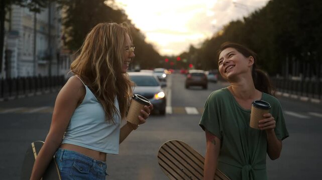 Close-up portrait of two laughing girl friends drinking coffee on a city street. Pretty smiling teenagers are hanging out enjoy time together with a skateboard and happy vibes