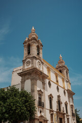 Baroque church facade with twin bell towers and clock faces under a clear blue summer sky in Algarve, Portugal.