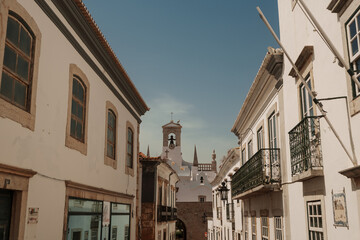 Algarve, Portugal – 2025.06.28 – A narrow street lined with whitewashed buildings leading to a church bell tower on a sunny day.