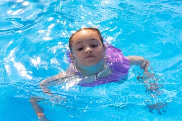 A young girl with dark hair joyfully swimming in a bright blue pool wearing a purple floatation...