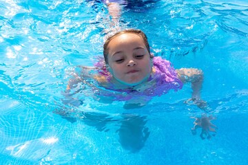 A young girl of Hispanic descent swims joyfully in a crystal-clear pool, showcasing the fun of...