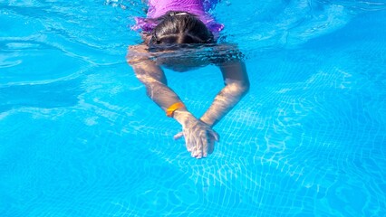A young girl with long hair swims gracefully underwater in a bright blue pool, showcasing her...