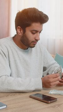 Young Indian man holds cash in dollars, thoughtfully plans expenses using calculator on smartphone, organizing money into separate piles on table in living room at home, managing finances carefully.