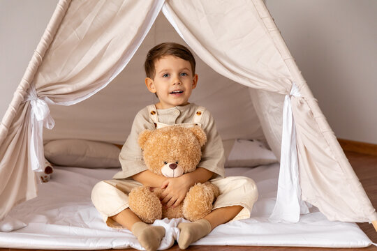 Happy 5 years old little boy sitting in a wigwam tent, hugging soft toy teddy bear. Children's room, childhood, games, imagination. High quality photo