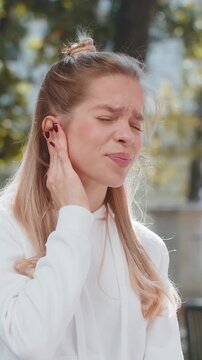 Earache. Sick Caucasian young woman suffers from ear pain sitting on chair in city street. Upset girl teenager touches her ear. She has furuncle, shingles, eczema, ear diseases, or otitis media.