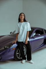 A studio photograph of a young woman standing next to a purple Porsche 911 sports car. The subject wears an oversized white t-shirt, loose black pants, and white sneakers. 