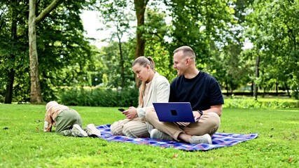 A young family couple works outdoors with a laptop while their child plays nearby. A happy family couple with a child are relaxing outdoors and working remotely on a laptop. - Powered by Adobe