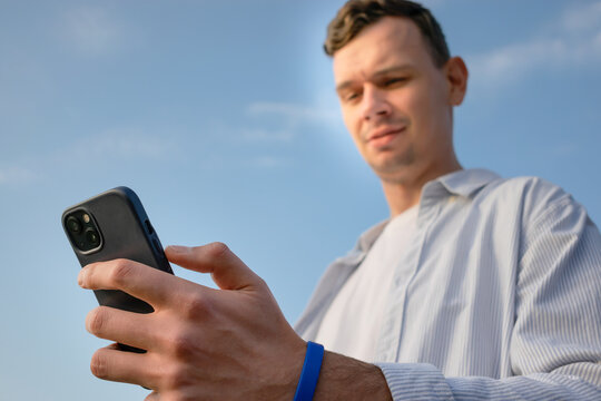 Young man holding smartphone outdoors against blue sky, close-up hand with phone, modern lifestyle.