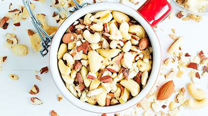 Overhead shot of a red-rimmed white bowl filled with a mix of chopped nuts, including cashews, almonds, and others, scattered on a white surface