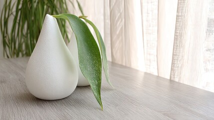 Two matte-white, pear-shaped vases sit on a light wood surface near a window, one holding a single, vibrant green plant sprig.  Soft, diffused light illuminates the scene
