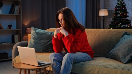 Woman celebrating new years eve at cozy home, woman resting sitting on the sofa, working on laptop, looking at screen surfing online, concentrated expression.