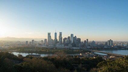 Fototapeta premium Cityscape view at dawn with skyscrapers, water, and trees under a clear blue sky in the morning light