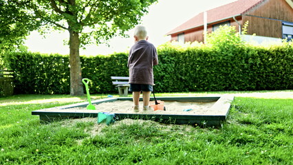 Toddler in a t-shirt and shorts on a walk on the playground. The boy plays in the sandbox, touching...
