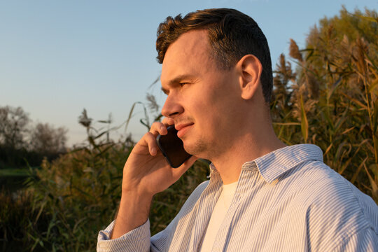 A young man talking on a smartphone outdoors during golden hour, standing near tall grass and enjoying warm sunlight.