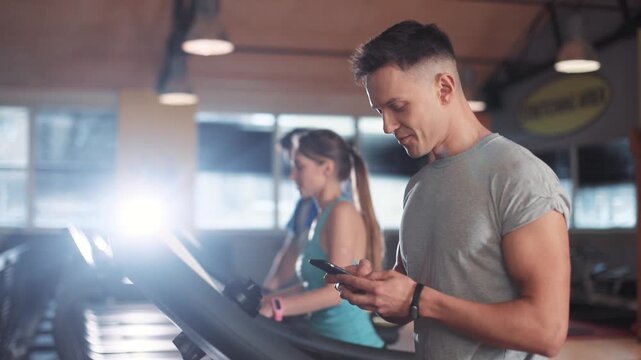 Smile young man use phone on a treadmill in gym sportsman sport fitness internet face technology fit lifestyle male workout athlete sportswear networking body exercise healthy mobile smartphone