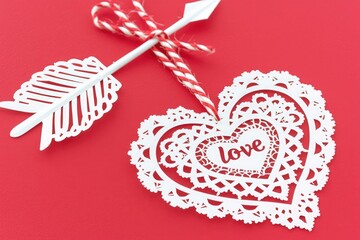 White paper cutout arrow with heart-shaped tip and love text beside heart-shaped lace doily on red background for romantic festive design