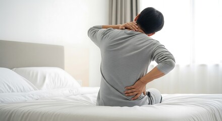 A man sits on a bed, holding his neck and lower back, indicating pain. He appears to be experiencing discomfort or stiffness in his upper and lower body. The setting suggests a bedroom.