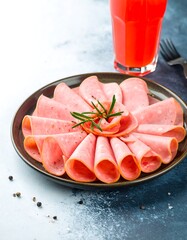 Thinly sliced deli meat arranged on a dark plate, garnished with rosemary, next to a glass of juice