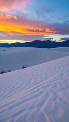 Vibrant sunset colors paint the sky above a vast expanse of white sand dunes, subtly textured by wind patterns
