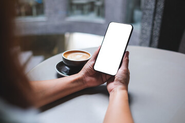 Mockup image of a woman holding and using mobile phone with blank desktop screen in cafe