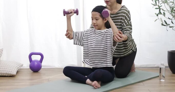 A young girl is training with dumbbells, with her mother helping her. Fit young people lift dumbbells and weights in a home, exercising for a healthy lifestyle concept