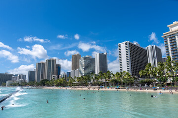 Naklejka premium Beachfront city skyline view with bathers on the sand and swimming in the sea with buildings and a beautiful blue sky with scattered clouds above.