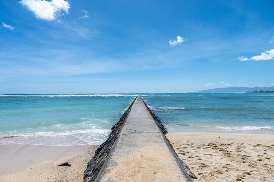 Ocean pier on a serene beach, blue sky overhead with fluffy clouds. A concrete pathway extends into the calming blue ocean.
