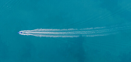 Aerial view of a speedboat cutting through clear turquoise sea, leaving a trail behind with anchored cargo ships in the distance. Ideal for marine transport, tourism, and coastal scenes.