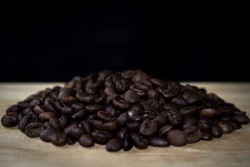 A pile of roasted coffee beans placed on a wooden surface with a black background. Perfect for food, beverage, cafe, and product design themes.