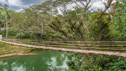 A rickety Bamboo Hanging Bridge over a tropical river. You can see the weaving of the path, the...