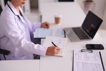 Adult Asian female doctor records medical patient data in office with laptop and documents healthcare tasks