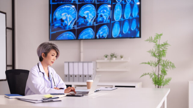 Asian Adult Female Doctor Analyzing Patient Medical Data on Laptop in Modern Clinic Office with MRI Scans