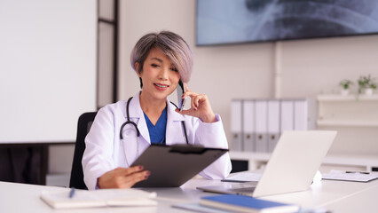 Asian adult female doctor communicates via phone in modern medical office for healthcare services