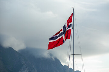 The Norwegian national flag fluttering from the mast of a yacht, with clouds and mountains in the background