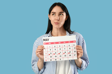 Thoughtful young woman with menstrual calendar on blue background
