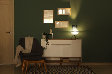 Interior of dark living room with glowing lamp on commode and armchair